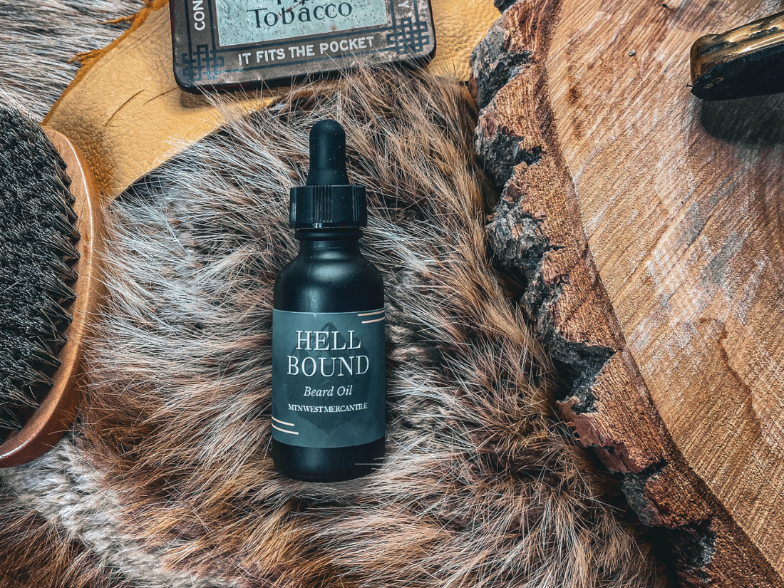 Mtn West Beard Co. Hell Bound beard oil bottle with dropper cap, styled on gray faux fur next to a vintage pipe tobacco tin, wooden beard comb, and a wood slice, with a leather strap in the background.