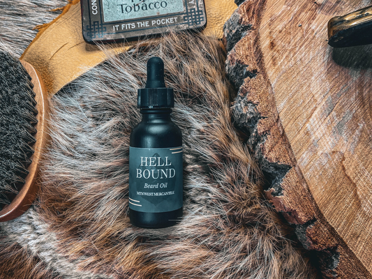 Mtn West Beard Co. Hell Bound beard oil bottle with dropper cap, styled on gray faux fur next to a vintage pipe tobacco tin, wooden beard comb, and a wood slice, with a leather strap in the background.