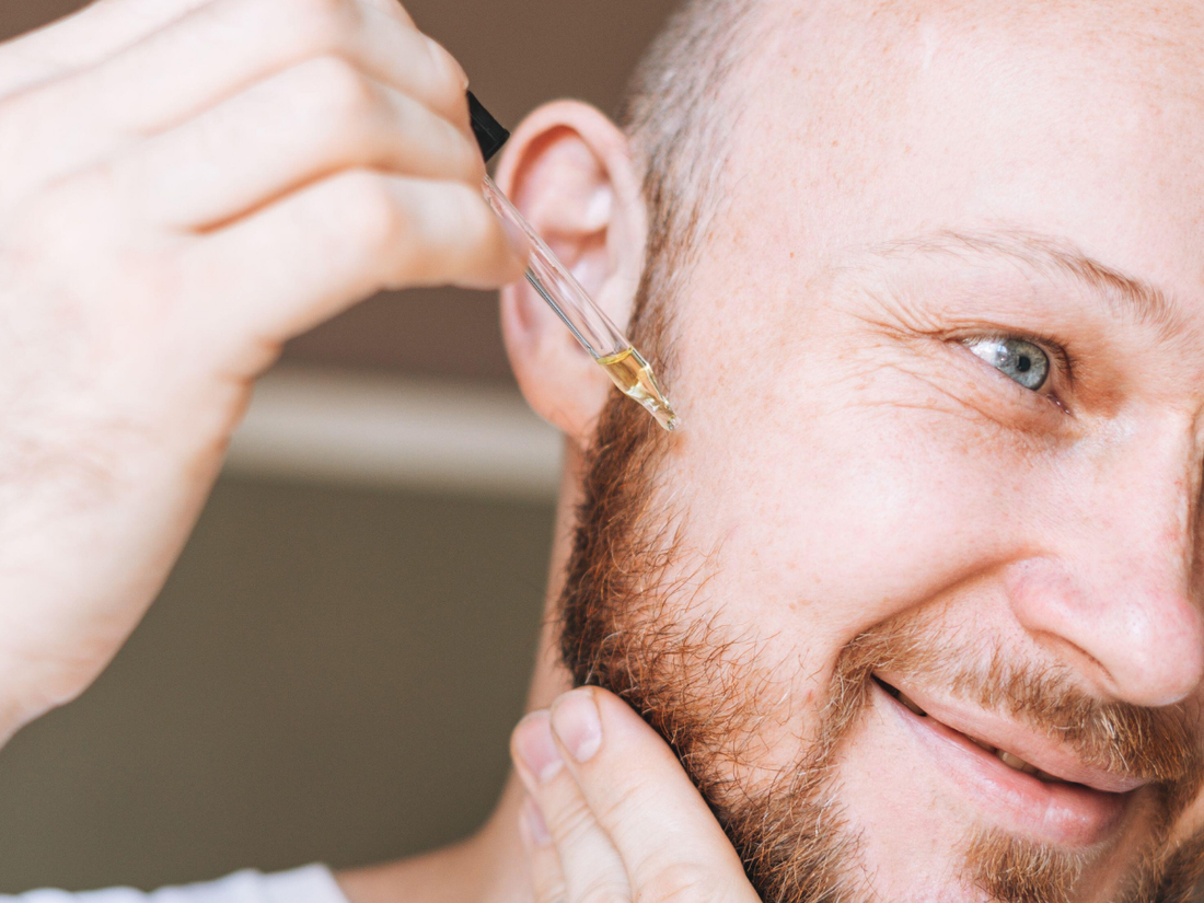 Close-up of a smiling red-bearded man applying beard oil from a dropper directly onto his beard near the jawline.